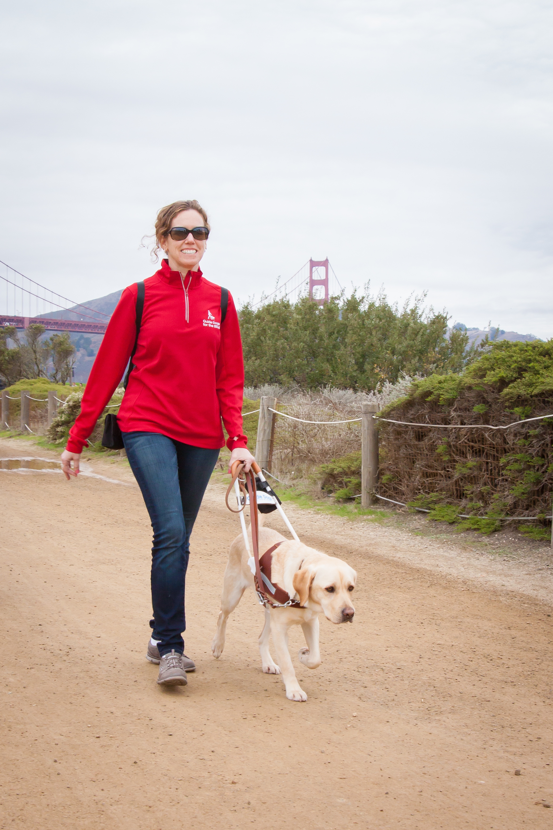 In this photo, Jane walks with her yellow Lab guide dog, Pilaf, along a coastal trail with the Golden Gate Bridge in the background. Jane smiles while looking forward and Pilaf looks focused on their destination.