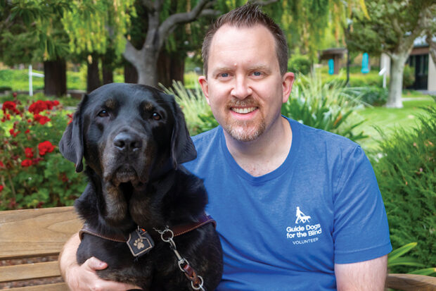 GDB guide dog team sits side by side on a bench. The client has their arm around the black Lab guide dog's shoulder.