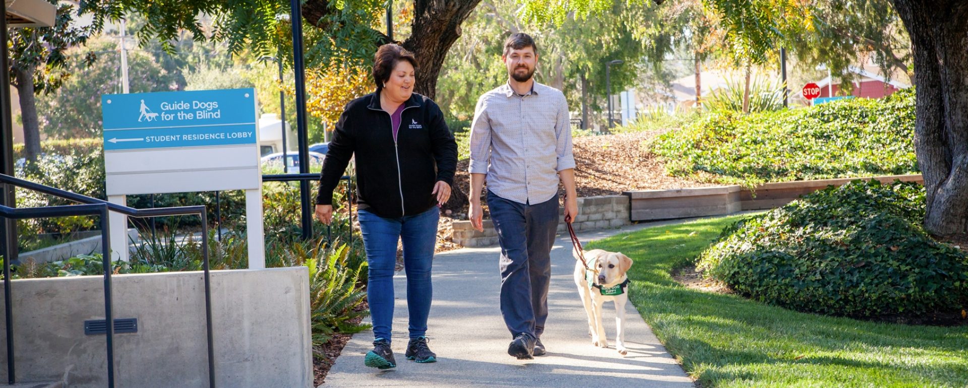 A woman and a man walking side by side down a path. The man is walking with a yellow Lab guide dog puppy wearing its green puppy coat.