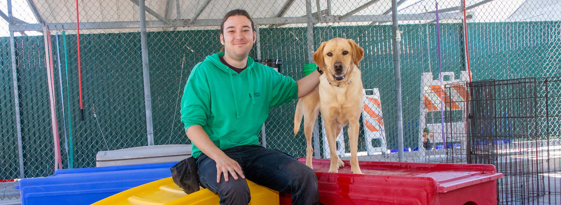 A man seated beside a yellow Lab guide dog in the dog play run.