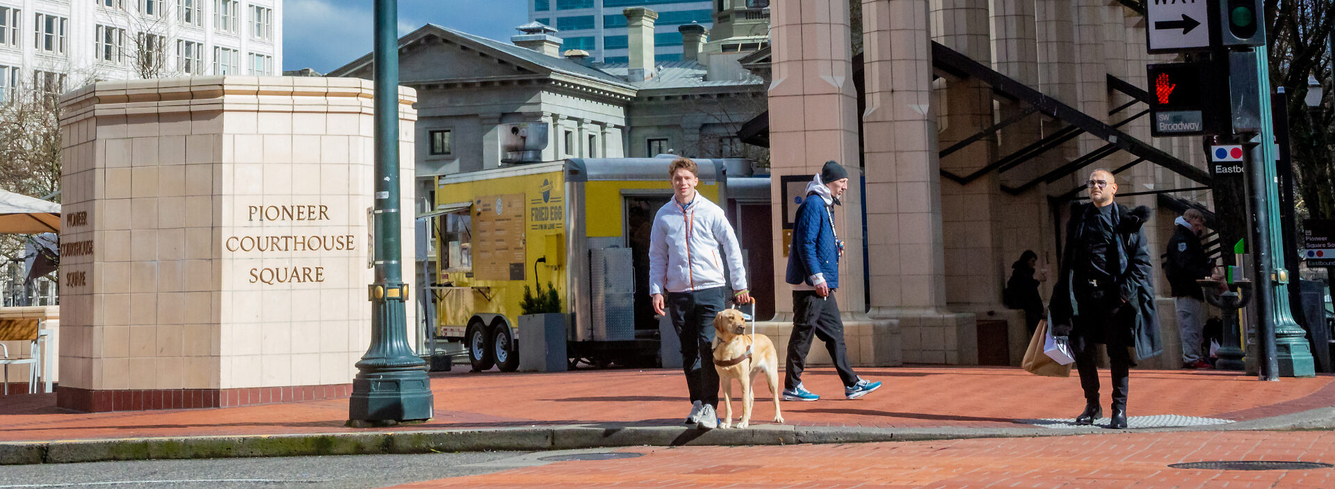 An instructor stands at a crosswalk with a yellow Lab guide dog.