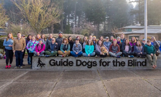 A group of instructors and apprentices stand together by a Guide Dogs for the Blind sign.