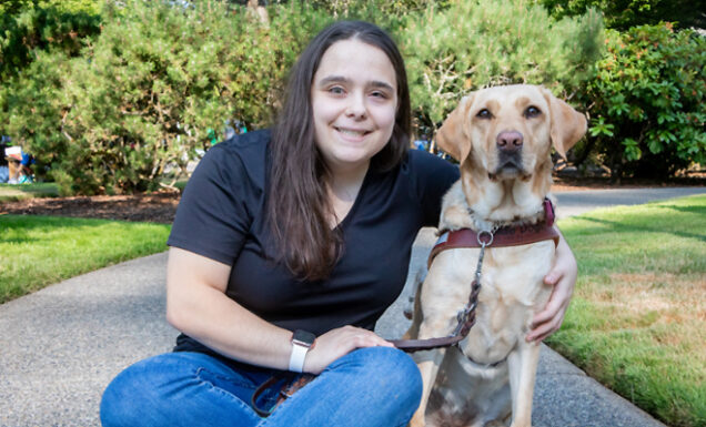 A staff member and her yellow Lab guide dog seated together on a grass-lined trail.
