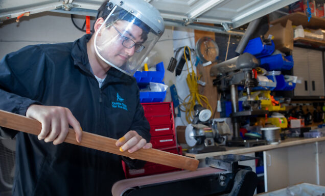 A man wearing protective gear sands down the edge of a piece of wood on a machine.