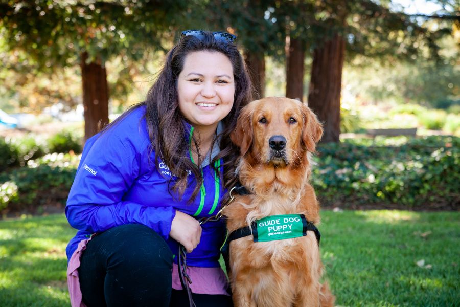 Devin seated next to a Golden Retriever guide dog puppy with a lawn and grove of trees in the background.