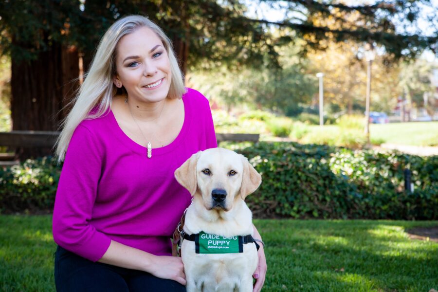 A photo of Alexandra kneeling on a green lawn next to a yellow Lab guide dog puppy. She is wearing a bright pink shirt and the puppy is wearing a green puppy-in-training jacket.