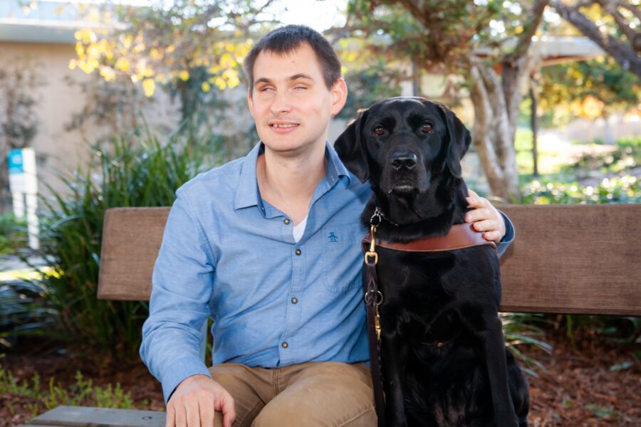A portrait of Jake sitting on a bench next to his black Lab guide dog.