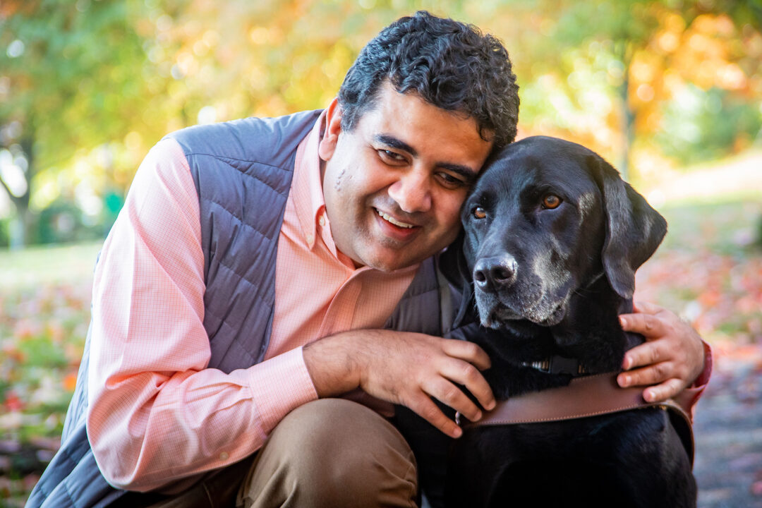 A smiling man kneels and embraces his black Lab guide dog.