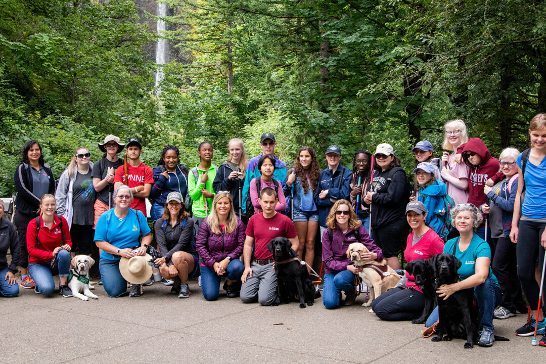 A large group of people and dogs, including teenagers with their white canes, in front of a picturesque waterfall in a lush forest.