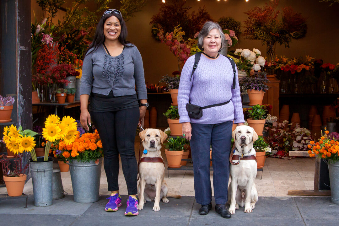 Two women with their guide dogs stand in front of a colorful flower shop.