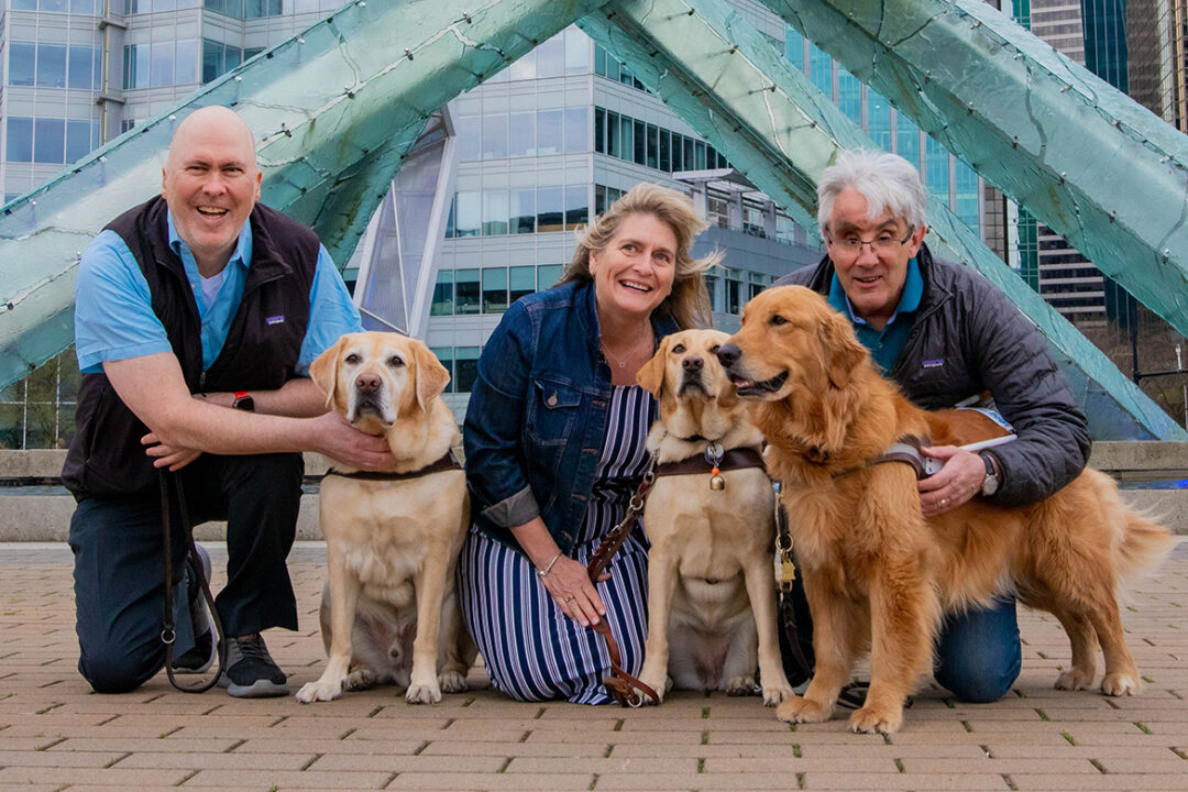 Three people kneel next to their guide dogs in Jack Poole Plaza in Vancouver, British Columbia.