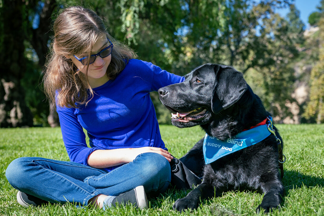 A teen girl sits next to her black Lab K9 Buddy on a green lawn.