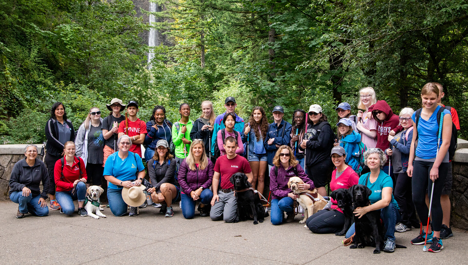 A large group of people and dogs, including teenagers with their white canes, in front of a picturesque waterfall in a lush forest.