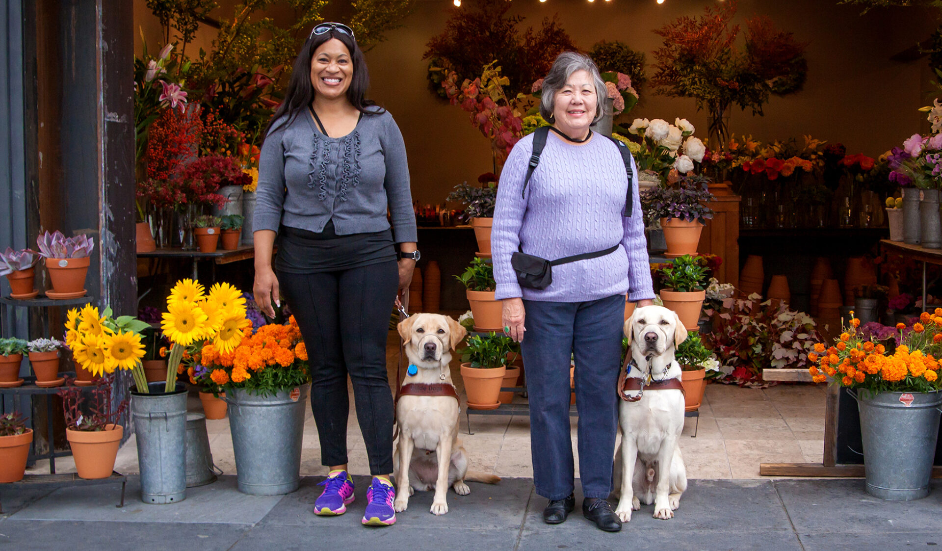 Two women with their guide dogs stand in front of a colorful flower shop.