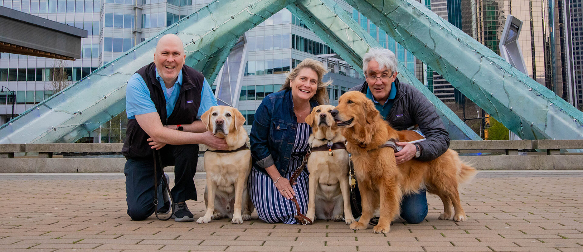 Three people kneel next to their guide dogs in Jack Poole Plaza in Vancouver, British Columbia.