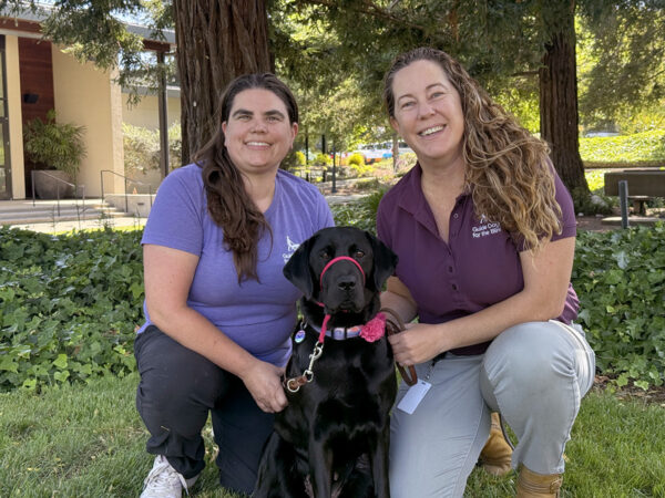 Two women kneel on a lawn with a black Lab.