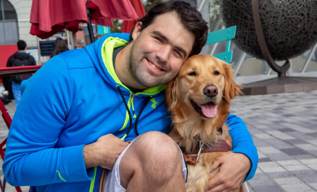 A smiling man kneels and embraces his grinning Golden Retriever guide dog.