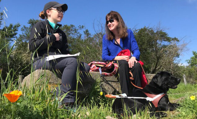 Sitting in an outdoor field of wildflowers, a guide dog mobility instructor chats with a woman whose guide dog lies at her feet.