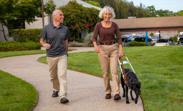 A guide dog mobility instructor walks on a path with a client and her new guide dog on GDB's California campus.