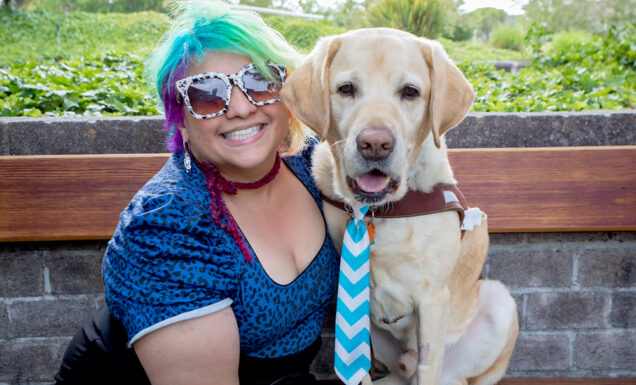 A person with bright multi-colored hair sits next to a smiling yellow Lab guide dog wearing a striped tie.