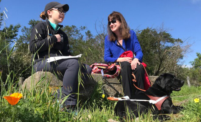 Sitting in an outdoor field of wildflowers, a guide dog mobility instructor chats with a woman whose guide dog lies at her feet.