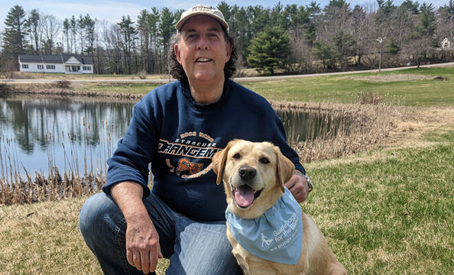 With a picturesque pond, farm house, and forest in the background, a man kneels next to a yellow Lab wearing a blue K9 Buddy scarf.