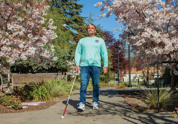 A man walks along a tree-lined path on a sunny day, using a cane for guidance.