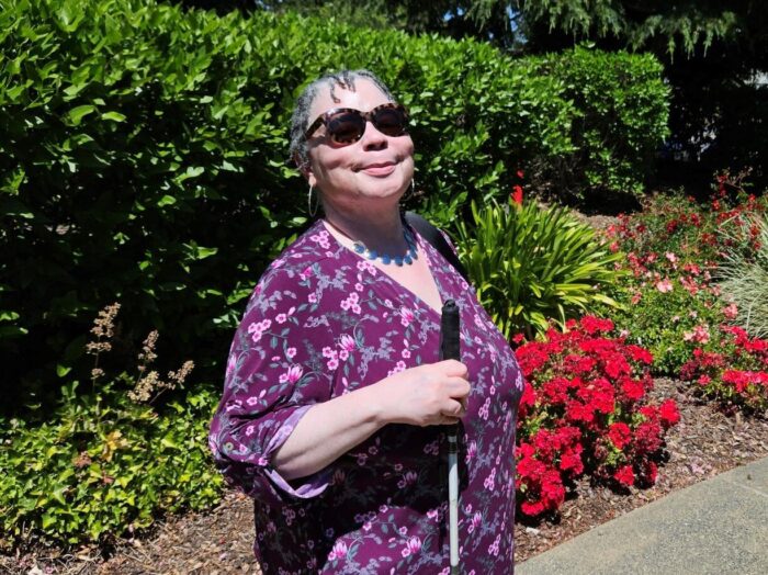 A woman holding a cane smiles as she stands on a shrub and flower lined path.