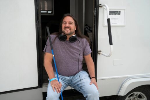 Joe is seated on the steps of a trailer on the set of a movie. He wears headphones around his neck and holds a cane in his right hand.