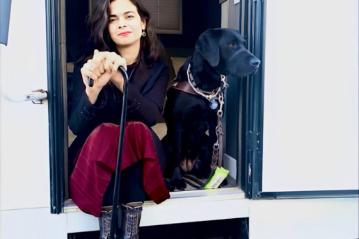 A woman sits in the open door of a trailer next to a black Lab guide dog.