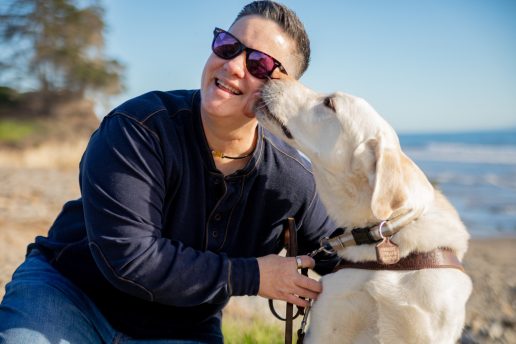 Cheri is seated beside her yellow Lab guide dog, Martinez, near the coast. Martinez lands a big kiss on her cheek while Cheri laughs.