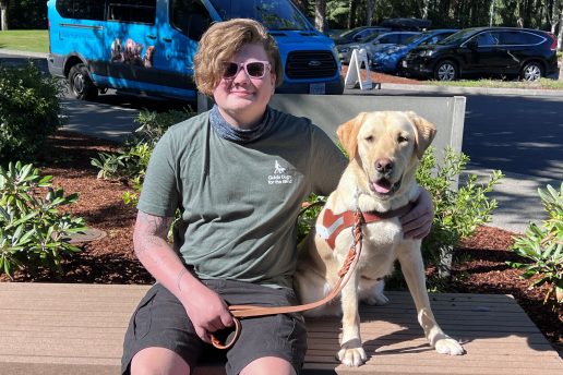 A young adult is seated beside a yellow Lab guide dog following a guided walk during Camp GDB.