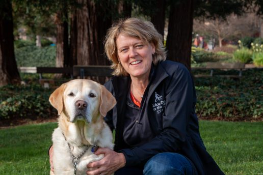 Dr. Kate kneels beside a yellow Lab. Kate is wearing a black Staff jacket and the dog looks seriously at the camera.