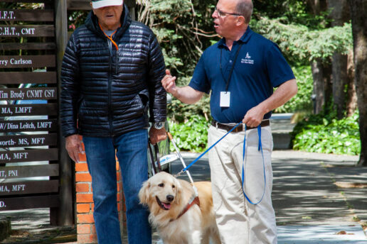Marc provides instruction at a crosswalk for a client during a guide dog walk. The Golden Retriever stands between the two men awaiting instructions.