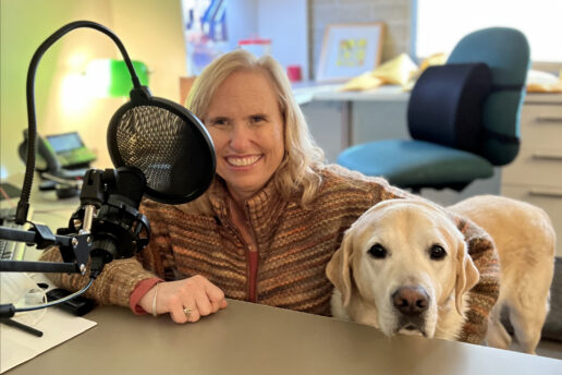 A woman sits at a desk behind a microphone with her guide dog by her side.