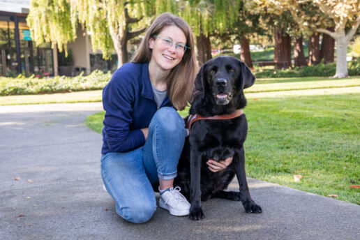 A woman kneels next to a black Lab on a walking path.