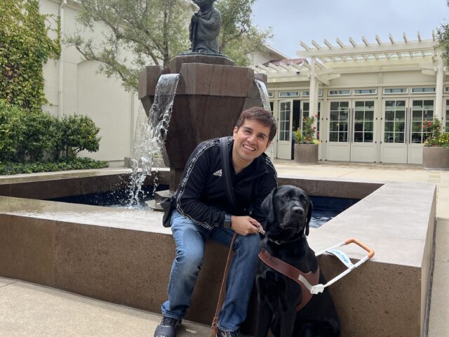Belo is seated beside a fountain with his black Lab guide dog, Limo by his side.