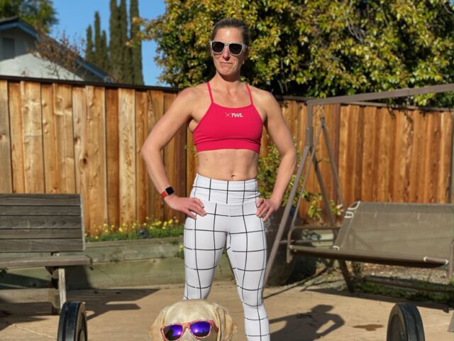 Kym stands confidently with her hands on her hips. In front of her is a large barbell loaded with heavy weights. In front of that is her retired yellow Lab guide dog, Citrus. Both Kym and Citrus wear matching sunglasses as the look into the camera.