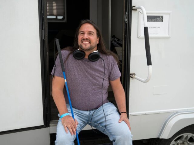 Joe is seated on the steps of a trailer on the set of a movie. He wears headphones around his neck and holds a cane in his right hand.