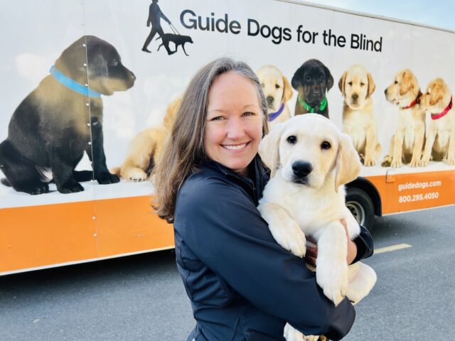 Sarah embraces a yellow Lab puppy. They are standing in front of the GDB Puppy Truck