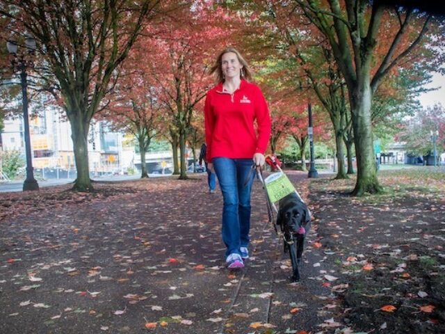 A woman in a red sweater walking with a black Lab guide dog down a tree-lined path.