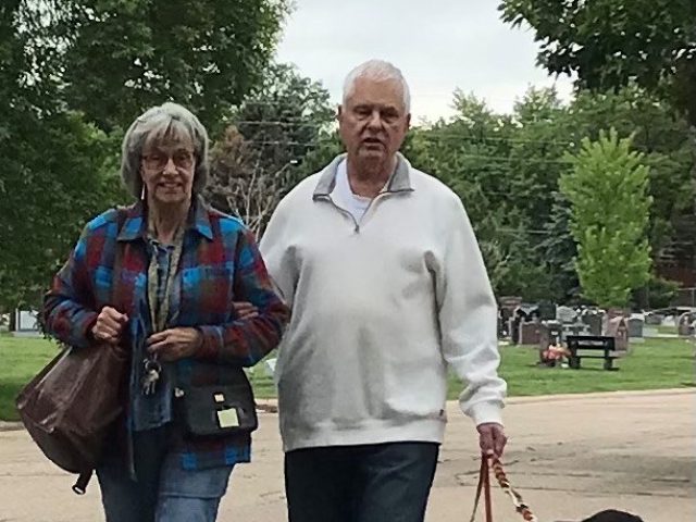 Steve walks beside his black Lab K9 Buddy, Bill. Steve holds a leash in one hand and his partner's arm in the other.