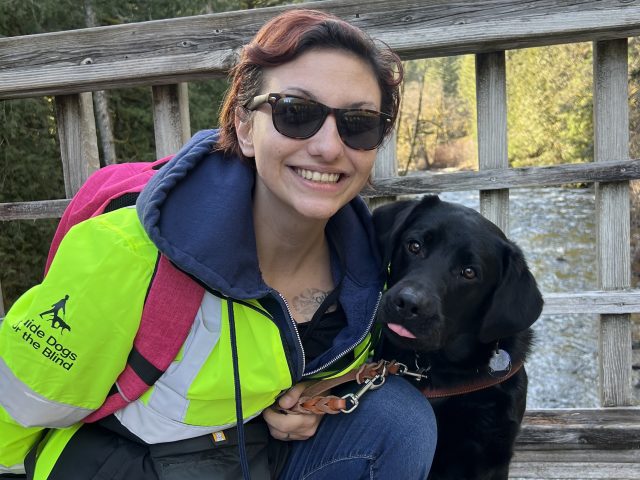 Brooke kneels beside her black Lab guide dog on a bridge. Brook is wearing her reflective GDB jacket and a red backpack. The dog looks curiously at the camera.