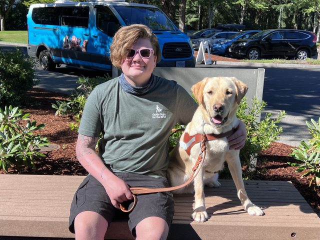 A young adult is seated beside a yellow Lab guide dog following a guided walk during Camp GDB.