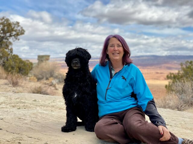 Jenna is seated beside her pet dog in front of a high desert vista.