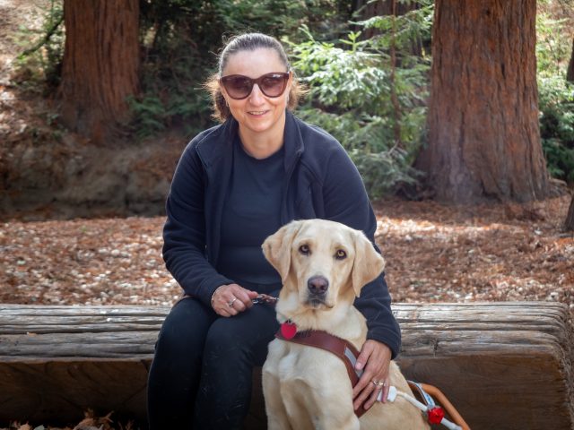 Kate sits on a log beside her yellow Lab guide dog. She rests one hand on the dog's shoulder and holds the leash with the other.