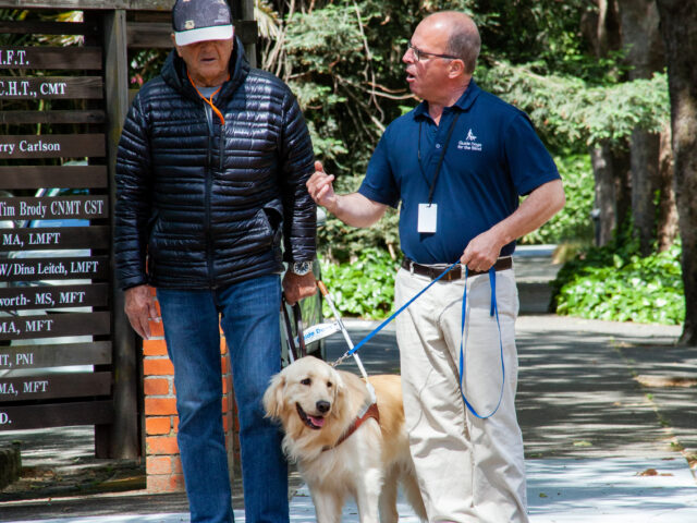 Marc provides instruction at a crosswalk for a client during a guide dog walk. The Golden Retriever stands between the two men awaiting instructions.