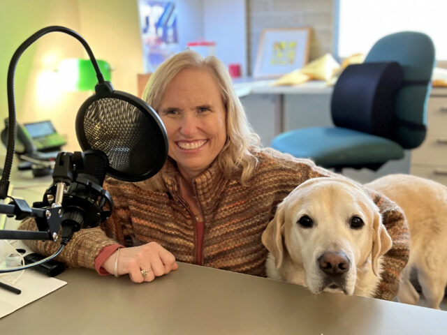A woman sits at a desk behind a microphone with her guide dog by her side.