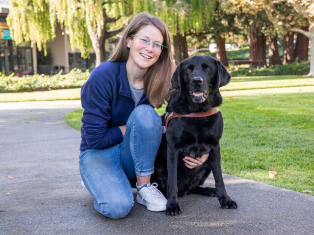 A woman kneels next to a black Lab on a walking path.