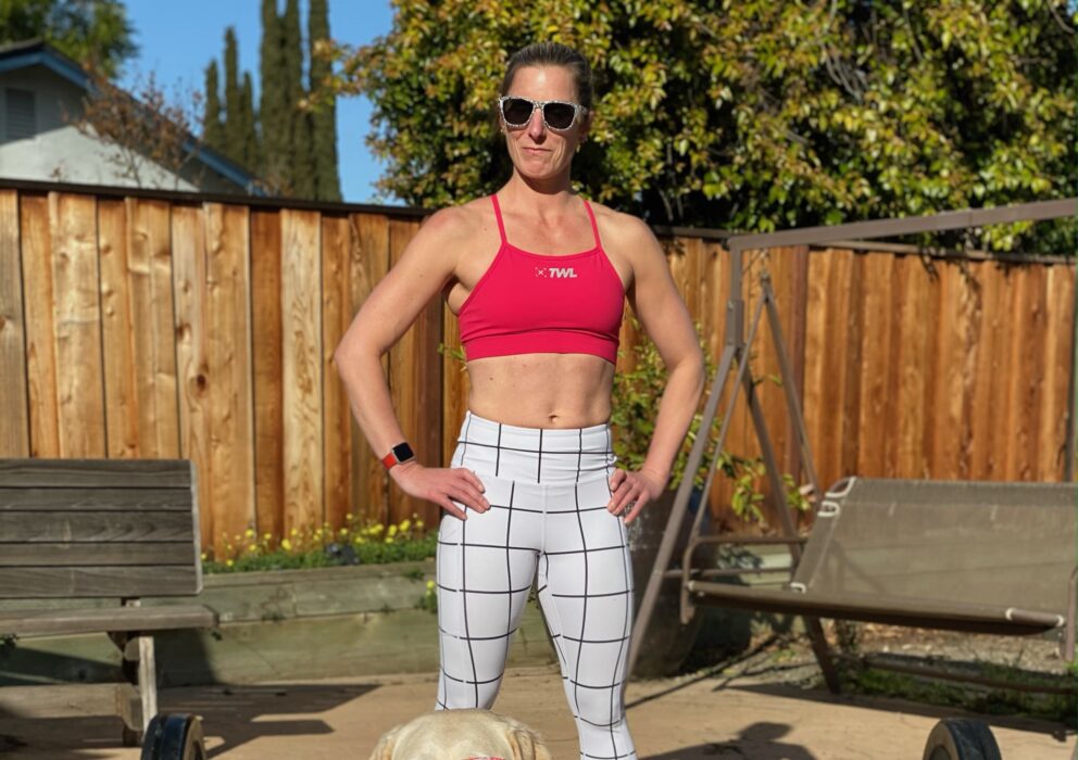 Kym stands confidently with her hands on her hips. In front of her is a large barbell loaded with heavy weights. In front of that is her retired yellow Lab guide dog, Citrus. Both Kym and Citrus wear matching sunglasses as the look into the camera.
